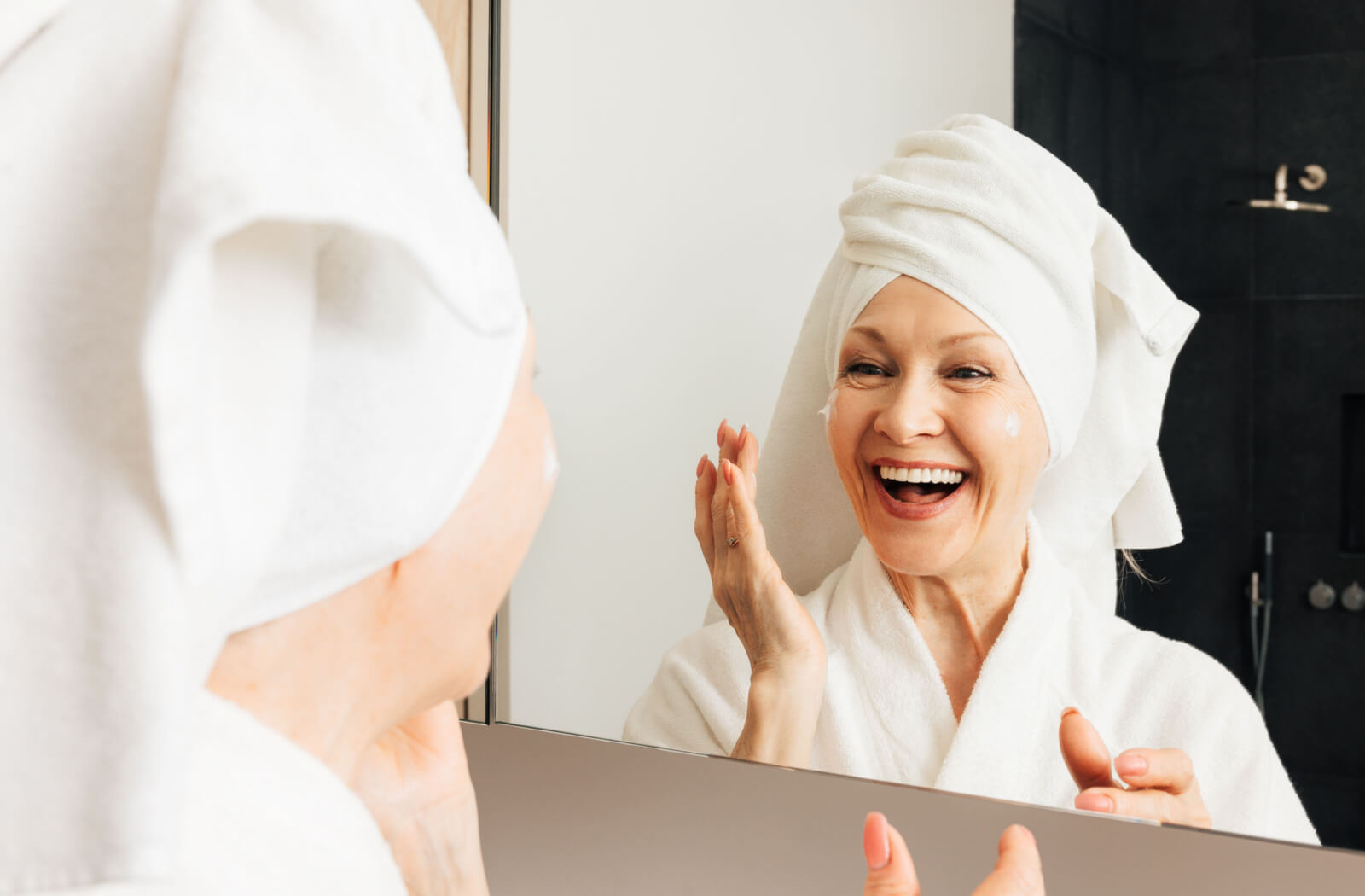 An older adult woman applies moisturizer to her face after her bath.