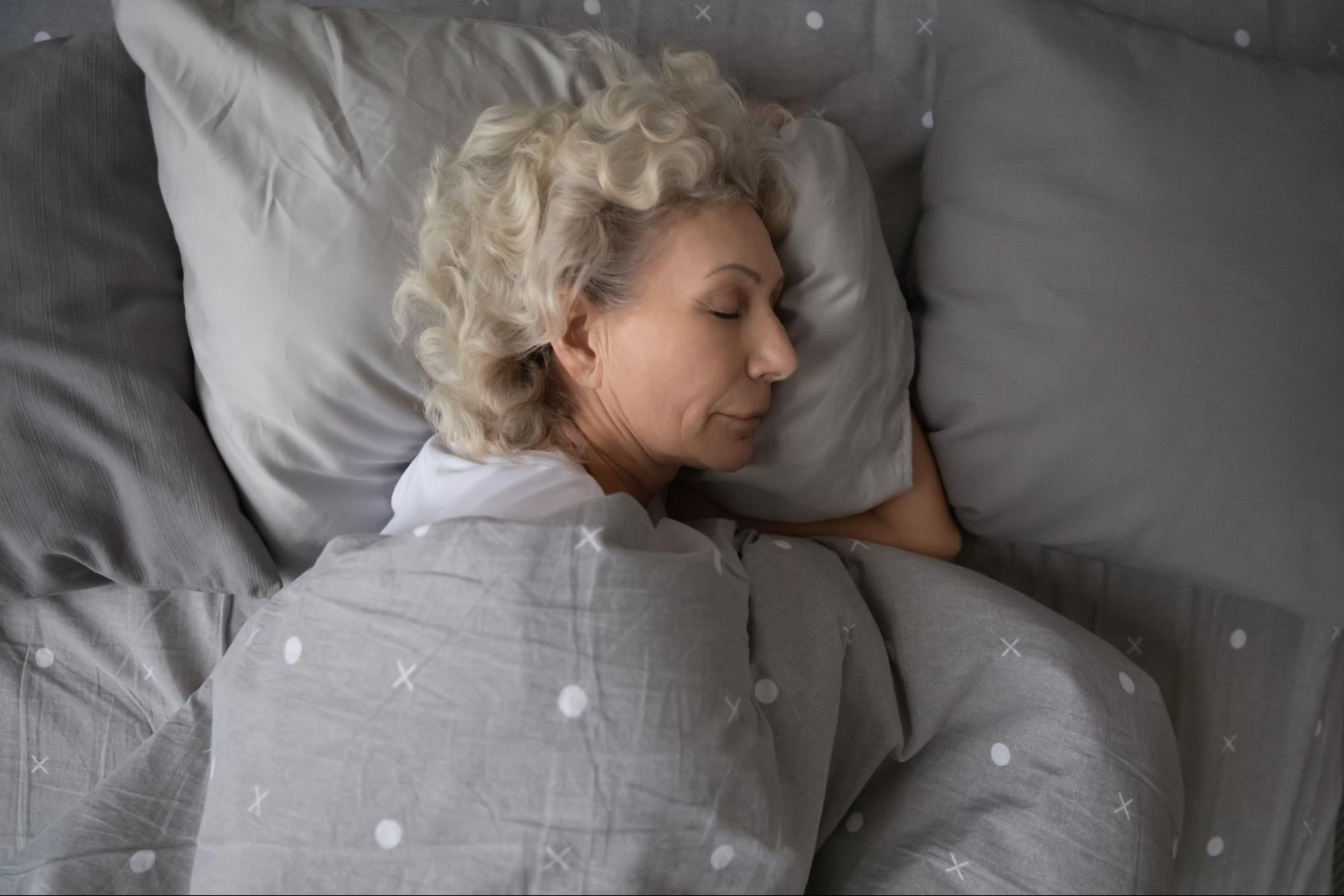 An overhead shot of a senior woman sleeping on a bed with gray blankets.