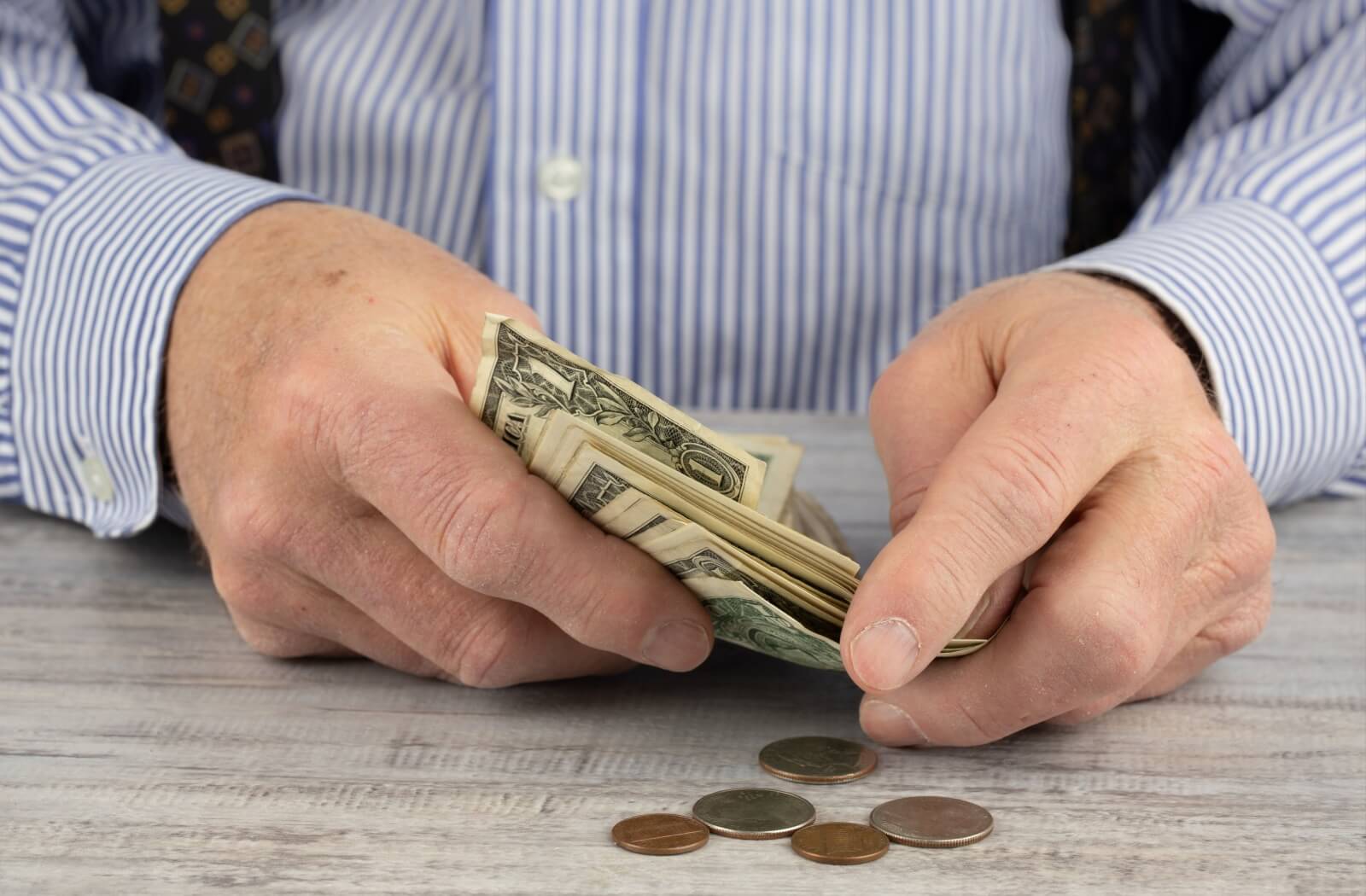 Close-up of an old man’s hands holding a wad of US bills, with some miscellaneous coins on the table in front of him