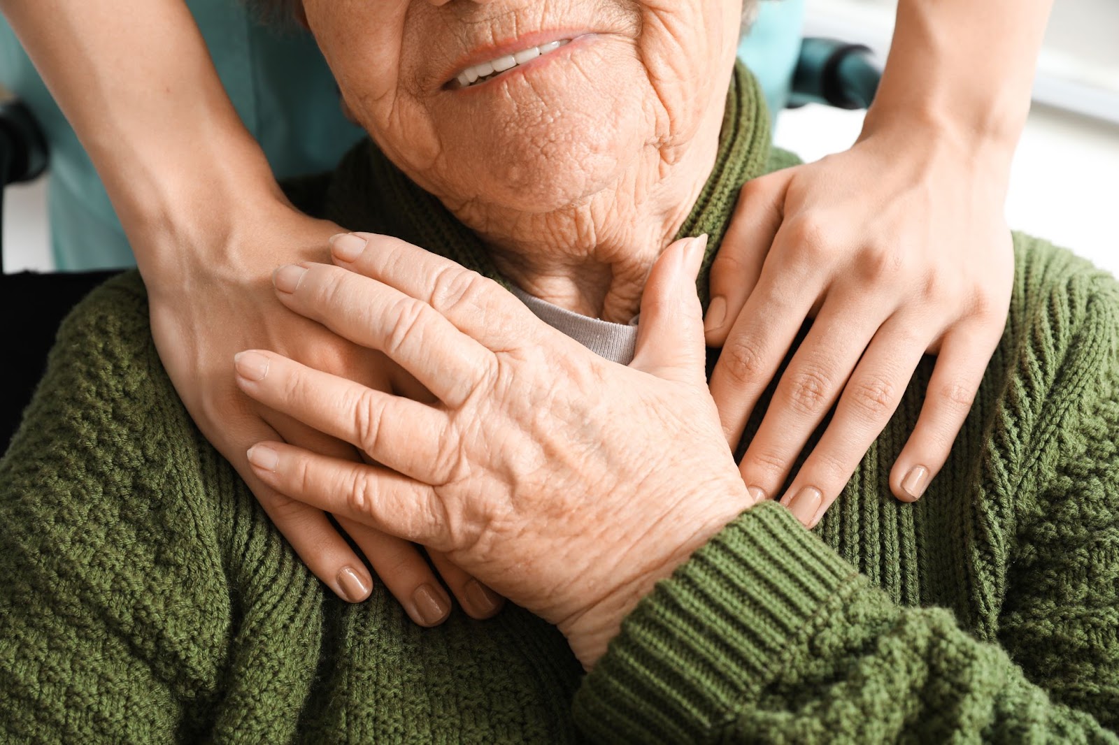 A young caregiver places their hands on an older adult's shoulders. The older adult reaches up to grasp the caregiver's hand.