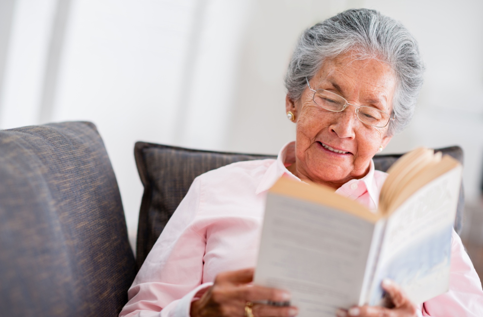 An older adult smiles while reclining on their couch and reading a white paperback novel