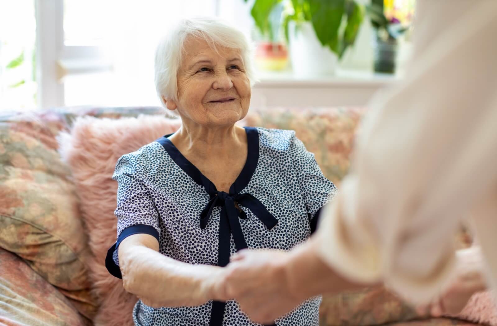 a senior woman sits on a couch, with a carer helping her up